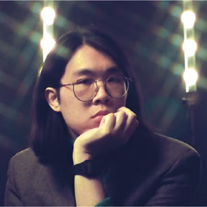 Portrait of Cameron Lew, chin on fist in round glasses, stage lights behind him.