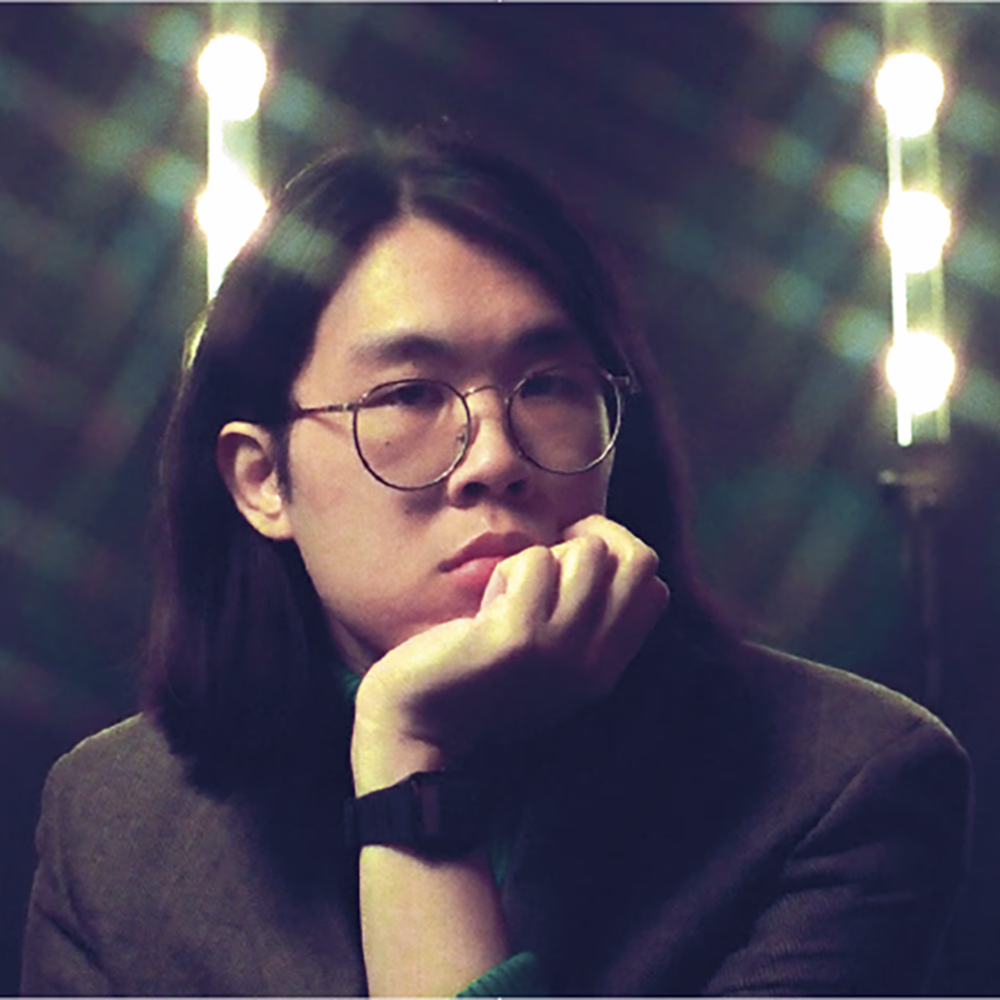 Portrait of Cameron Lew, chin on fist in round glasses, stage lights behind him.