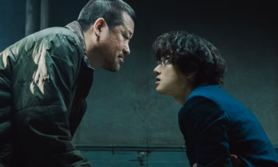 Two men lean face-to-face across an interrogation room in cold green light