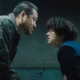 Two men lean face-to-face across an interrogation room in cold green light