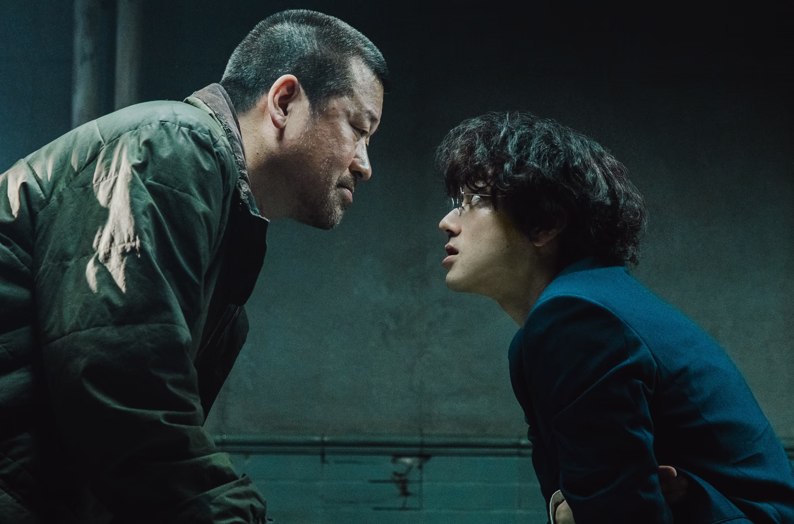 Two men lean face-to-face across an interrogation room in cold green light