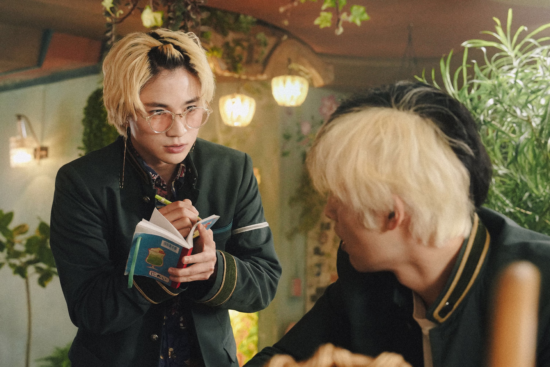 A blond student in tinted glasses jots in a tiny notebook inside a plant-filled cafe