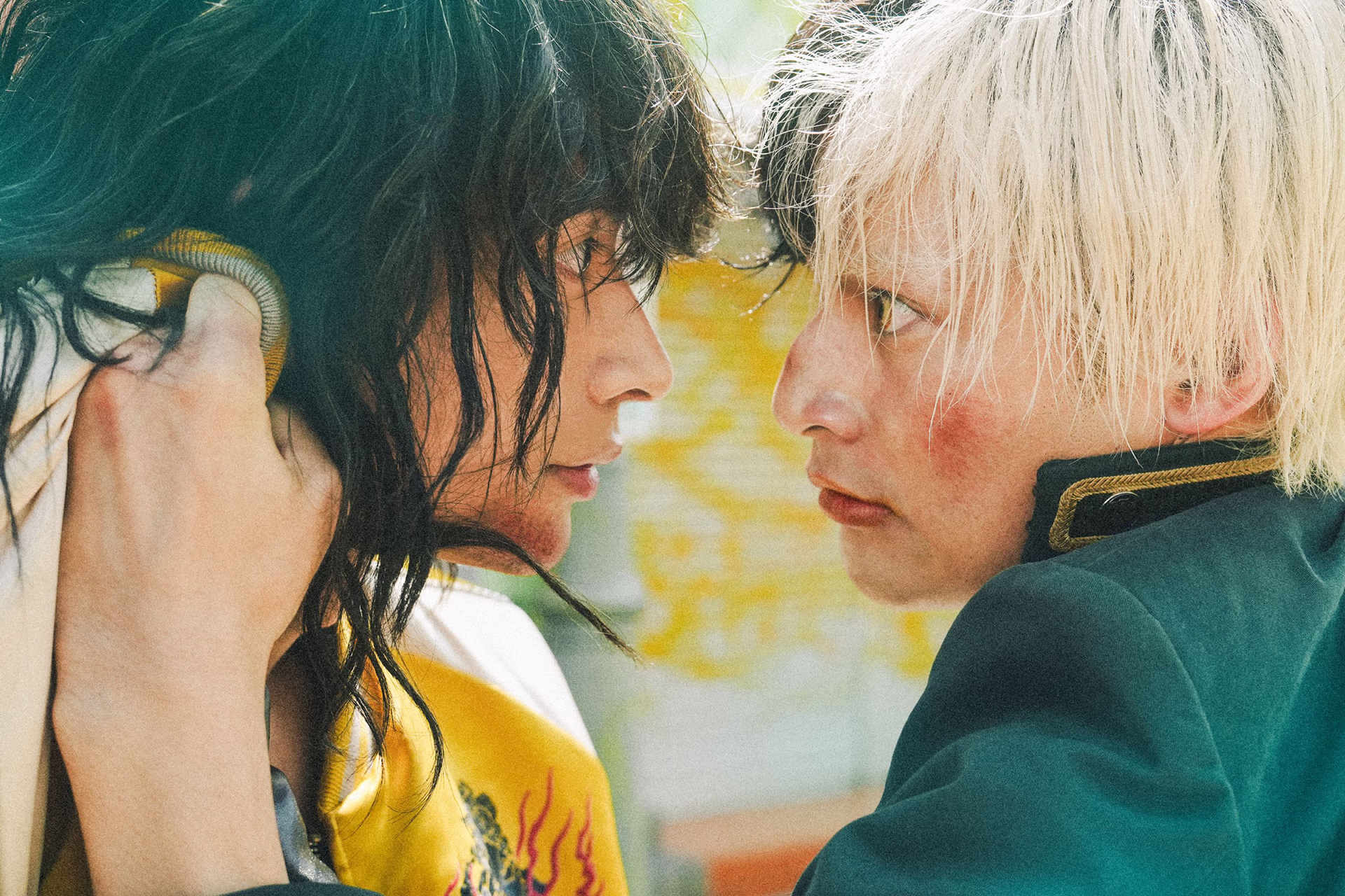 Two students grip each other nose-to-nose, one in a yellow souvenir jacket, one blond in green uniform
